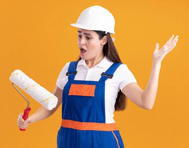 Scared young builder woman in uniform holding and looking at roller brush isolated on orange wall