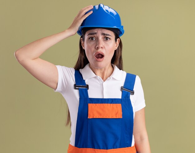 オリーブグリーンの壁に分離された頭に手を置く制服を着た若いビルダーの女性