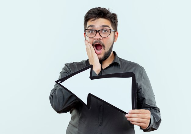 Scared young businessman wearing glasses holding direction mark isolated on white with copy space