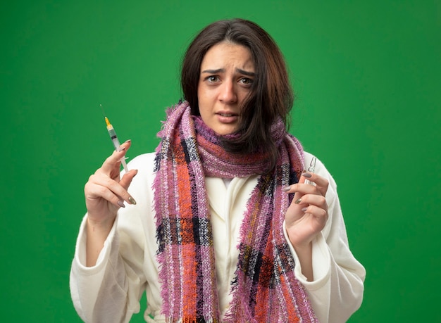 Испуганная молодая кавказская больная девушка в халате и шарфе держит ампулу и шприц, глядя в камеру, изолированную на зеленом фоне