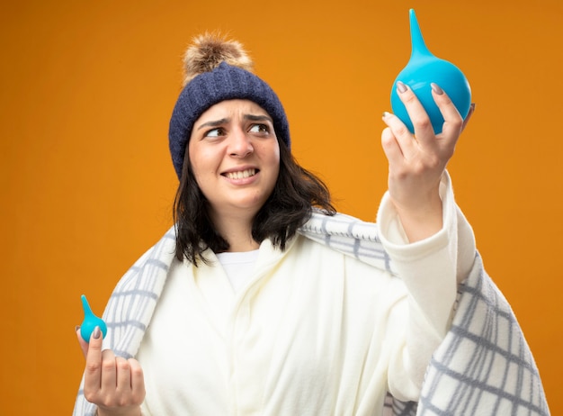 Scared young caucasian ill girl wearing robe winter hat wrapped in plaid holding enemas raising up big one looking at it isolated on orange background