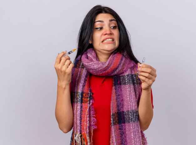 Scared young caucasian ill girl wearing scarf holding syringe and ampoule looking at ampoule isolated on white wall with copy space