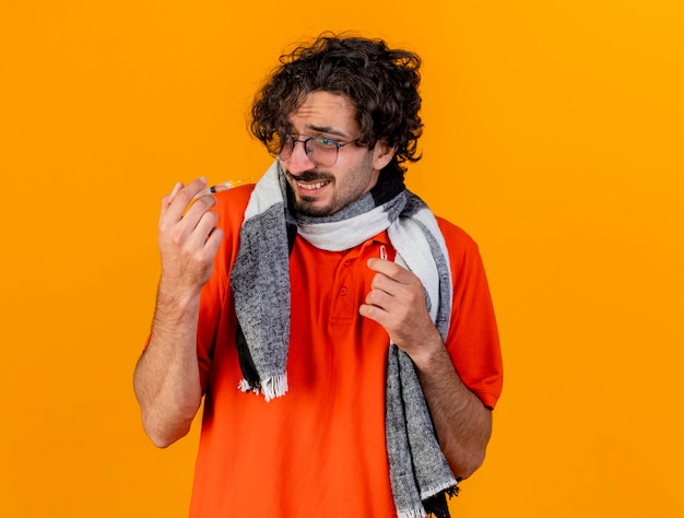 Scared young caucasian ill man wearing glasses and scarf holding syringe and ampoule looking at syringe isolated on orange wall with copy space