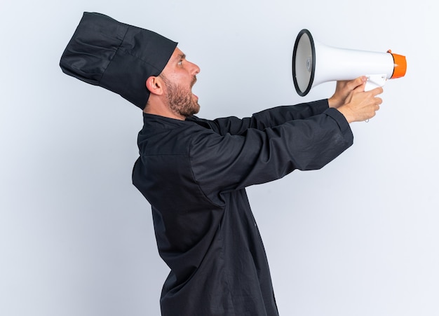 Scared young caucasian male cook in chef uniform and cap standing in profile view stretching out and looking at speaker isolated on white wall