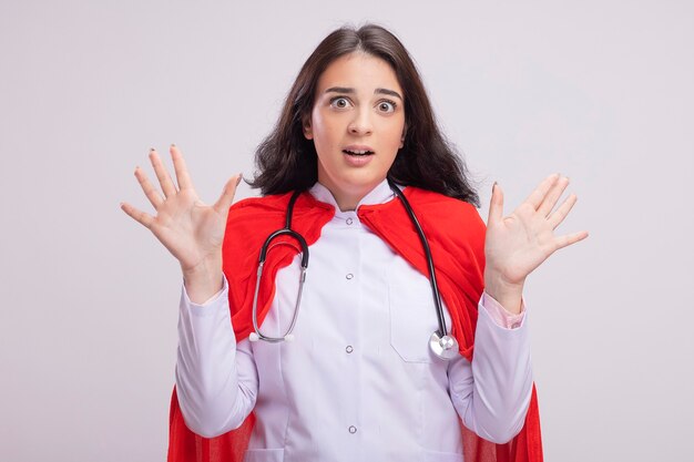 Scared young caucasian superhero girl wearing doctor uniform and stethoscope showing empty hands 