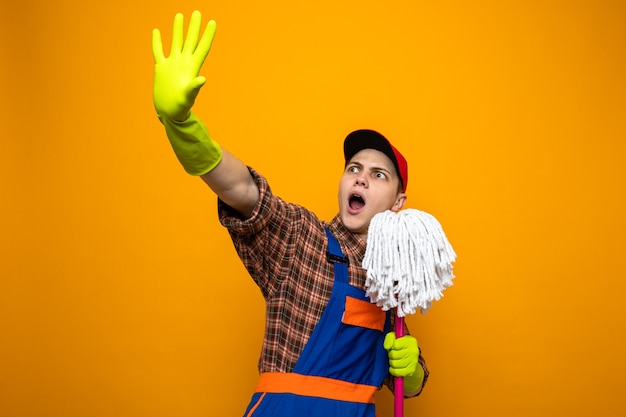 Foto gratuita giovane ragazzo delle pulizie spaventato che indossa uniforme e berretto con guanti che tengono mop