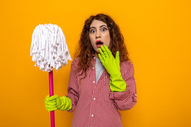 Scared young cleaning woman wearing gloves holding mop 