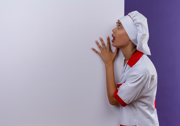 Scared young cook female wearing chef uniform holding white wall on isolated background with copy space