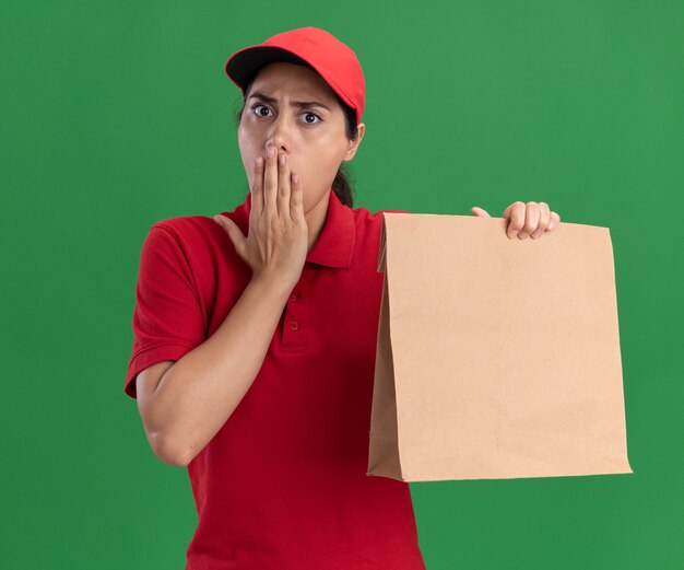 Scared young delivery girl wearing uniform and cap holding paper food package covered mouth with hand isolated on green wall