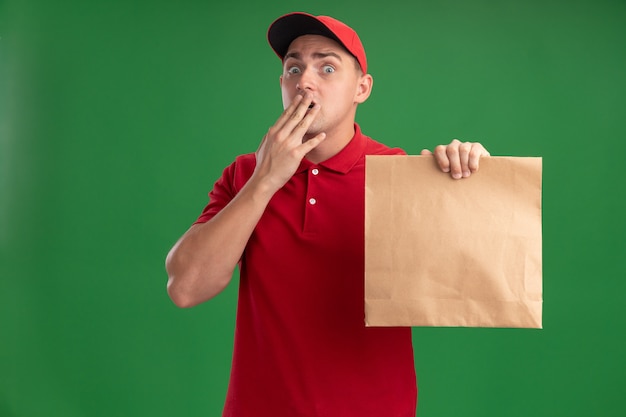 Spaventato il giovane fattorino che indossa l'uniforme e il cappuccio che tiene il pacchetto di cibo di carta coperto la bocca con la mano isolata sulla parete verde