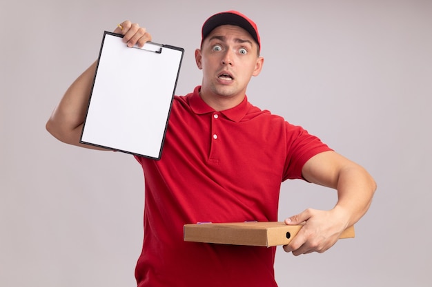 Испуганный молодой доставщик в униформе с кепкой, держащей буфер обмена с коробкой для пиццы, изолированной на белой стене