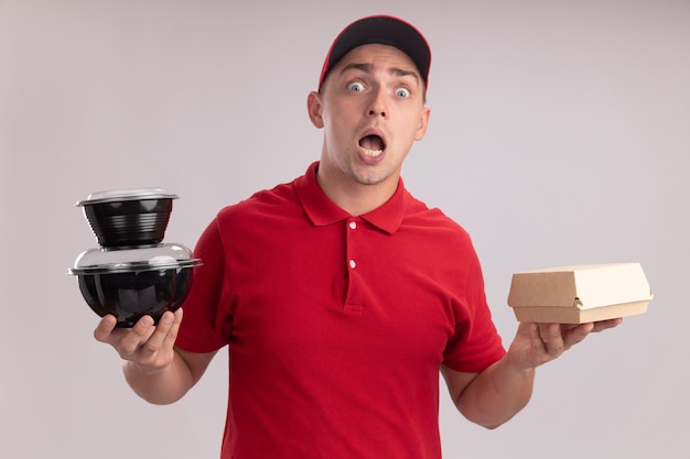 Foto gratuita giovane uomo di consegna spaventato che indossa l'uniforme con il cappuccio che tiene il pacchetto alimentare di carta con il contenitore per alimenti isolato sulla parete bianca