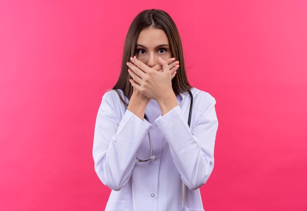 Испуганная молодая девушка-врач в медицинском халате со стетоскопом прикрыла рот на изолированном розовом фоне