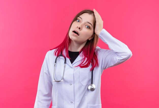 Испуганная молодая девушка-врач в медицинском халате со стетоскопом положила руку на голову на розовом изолированном фоне