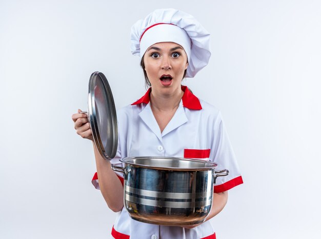 Scared young female cook wearing chef uniform holding saucepan with lid isolated on white background