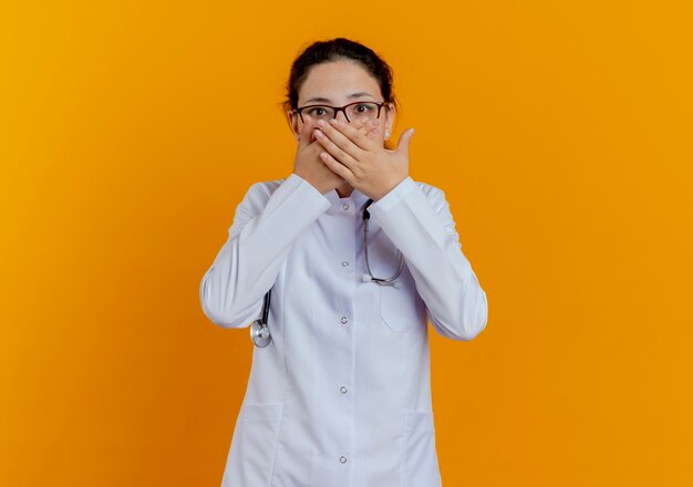 Scared young female doctor wearing medical robe and stethoscope with glasses covered with hands mouth isolated