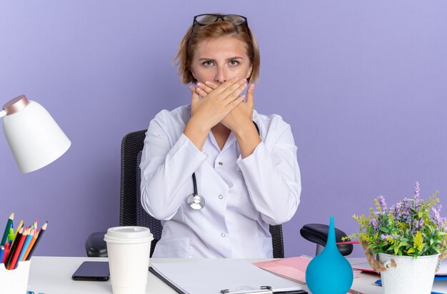 Scared young female doctor wearing medical robe with stethoscope and glasses sits at table with medical tools covered mouth with hands isolated on blue background