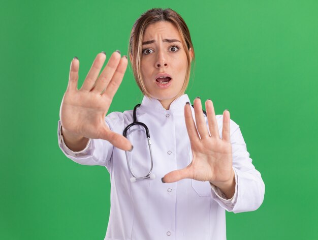 緑の壁に分離されたカメラに手を差し伸べる聴診器で医療用ローブを着た怖い若い女性医師