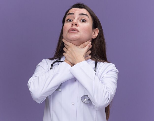 Scared young female doctor wearing medical robe with stethoscope pretends to choke herself with hands isolated on purple wall with copy space
