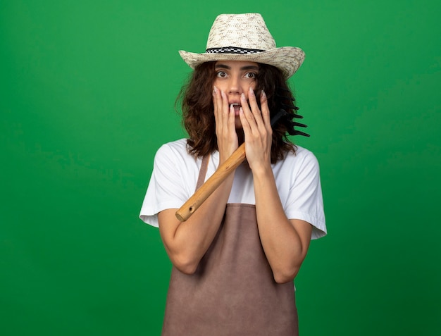 Scared young female gardener in uniform wearing gardening hat holding rake covered face with hands