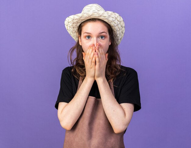 Scared young female gardener wearing gardening hat covered face isolated on blue wall