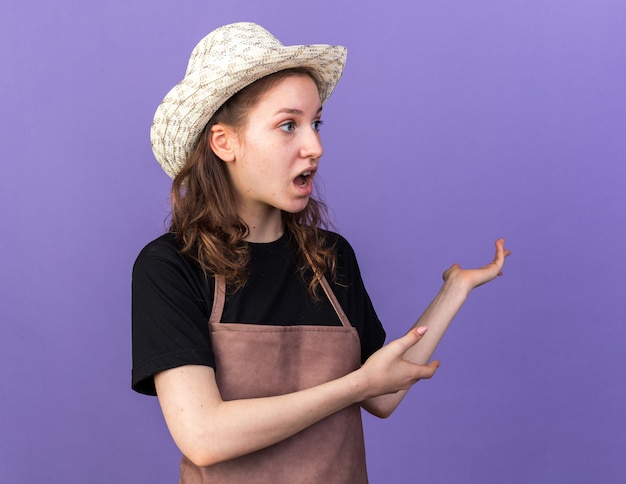 Scared young female gardener wearing gardening hat points at behind 