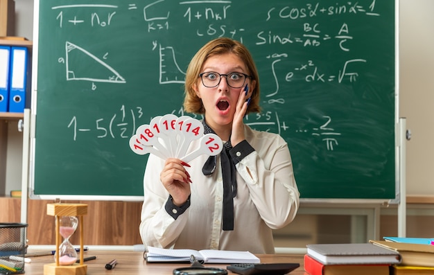 Scared young female teacher wearing glasses sits at table with school supplies holding number fans putting hand on cheek in classroom