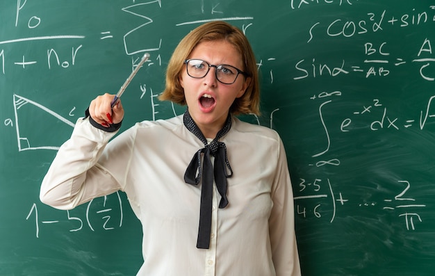 Scared young female teacher wearing glasses standing in front blackboard holding pointer stick in classroom