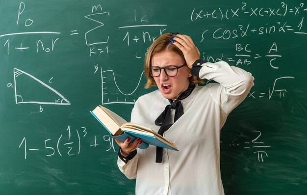 scared young female teacher wearing glasses standing in front blackboard reading book putting hand on head in classroom