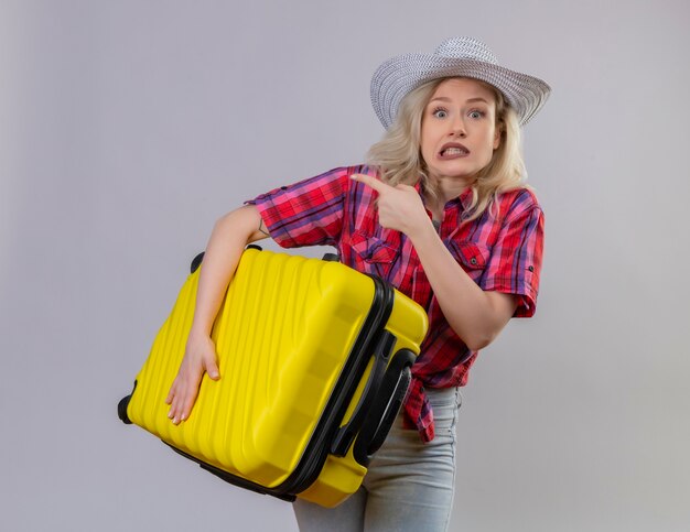 Scared young female traveler wearing red shirt in hat holding suitcase points to side on isolated white wall