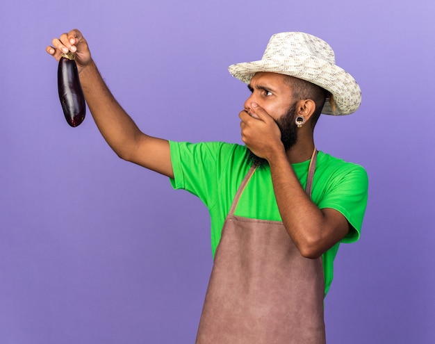 Foto gratuita giovane giardiniere spaventato afro-americano che indossa un cappello da giardinaggio che tiene e guarda la bocca coperta di melanzane con la mano