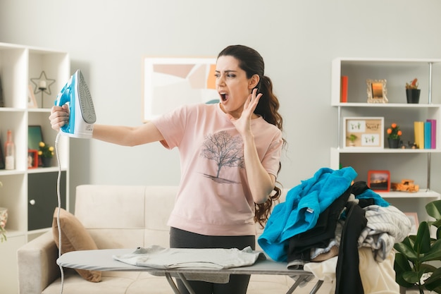 Scared young girl holding and looking at steam iron standing behind ironing board with clothes in living room