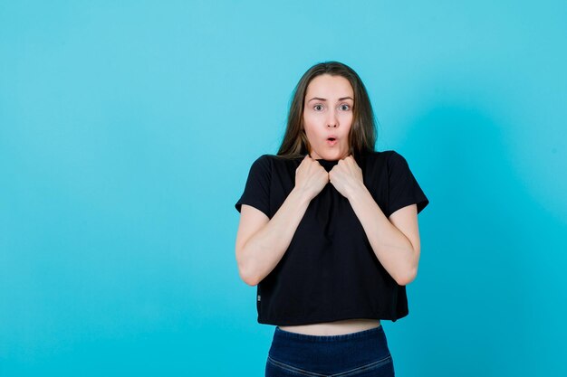 Scared young girl is holding fists on chest on blue background