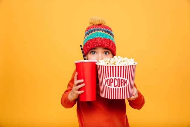 Scared Young girl in sweater and hat hiding behind the popcorn and plastic cup while looking at the camera over orange