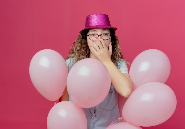 Scared young girl wearing glasses and pink hat standing among balloons and covered mouth isolated on pink wall