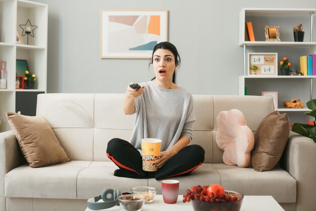 Испуганная молодая девушка с ведром попкорна, держащая пульт от телевизора, сидит на диване за журнальным столиком в гостиной