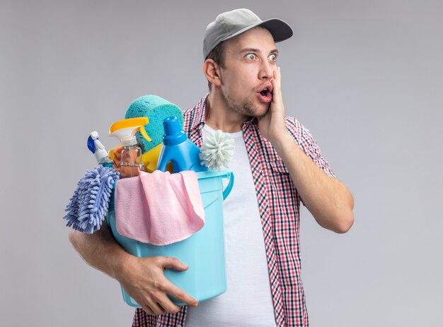 scared young guy cleaner wearing cap holding bucket with cleaning tools putting hand on cheek isolated on white wall