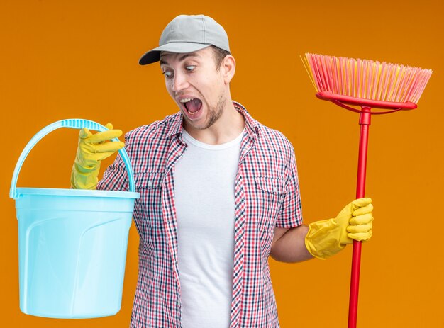 Scared young guy cleaner wearing cap with gloves holding bucket and mop isolated on orange wall