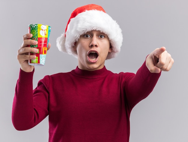 Scared young guy wearing christmas hat holding christmas cup points  isolated on white wall