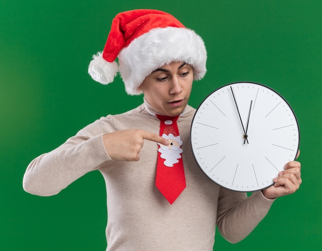 Scared young guy wearing christmas hat with tie holding and points at wall clock isolated on green wall