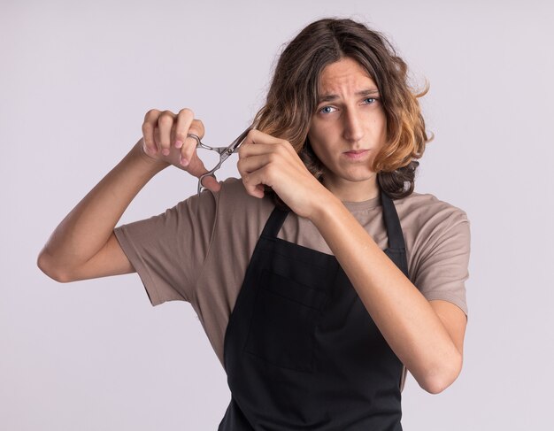 Scared young handsome barber wearing uniform cutting his own hair with scissors 