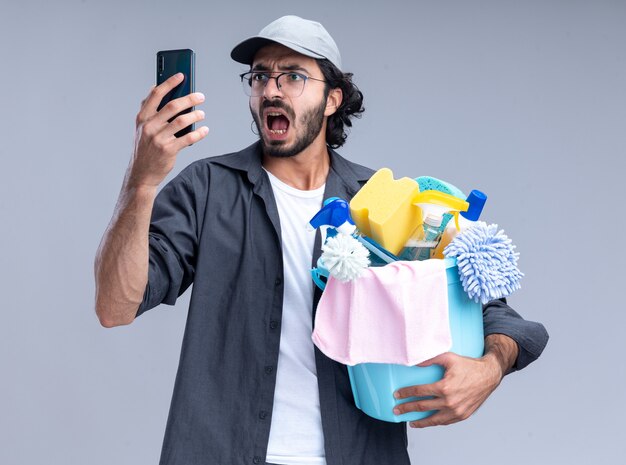 Испуганный молодой красивый уборщик в футболке и кепке, держащий ведро с чистящими инструментами и смотрящий на телефон в руке, изолированной на белой стене