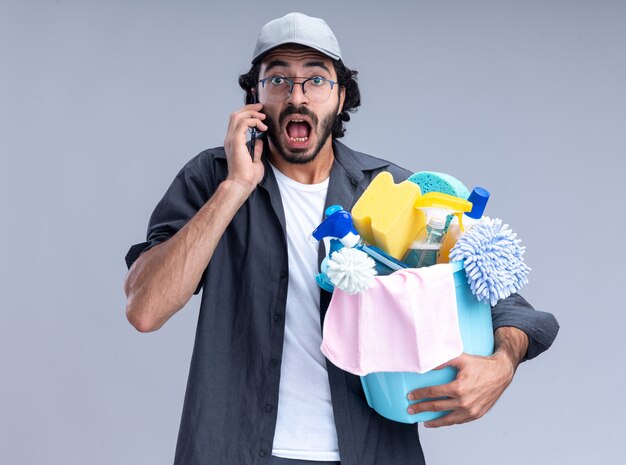 Scared young handsome cleaning guy wearing t-shirt and cap holding bucket of cleaning tools and speaks on phone isolated on white wall