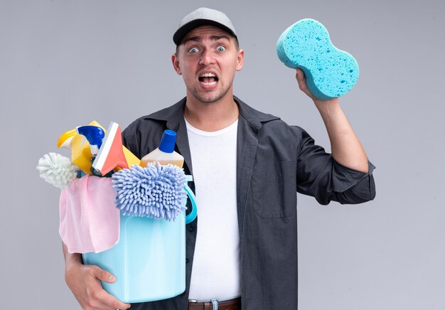 Испуганный молодой красивый уборщик в футболке и кепке, держащий ведро с чистящими инструментами и губкой, изолированным на белой стене