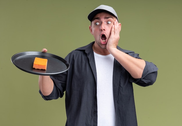 Free photo scared young handsome cleaning guy wearing t-shirt and cap holding and looking at sponge on tray putting hand on cheek isolated on olive green wall