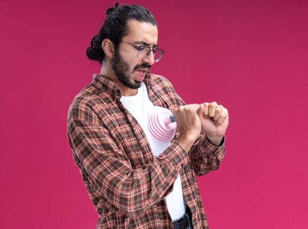 Scared young handsome cleaning guy wearing t-shirt putting plunger on heart isolated on pink wall