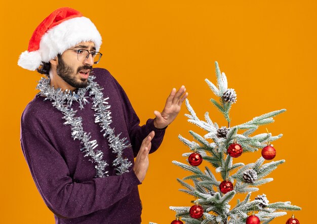 オレンジ色の背景で隔離のカメラで手を差し出して首に花輪とクリスマスの帽子をかぶってクリスマスツリーの近くに立っている怖い若いハンサムな男