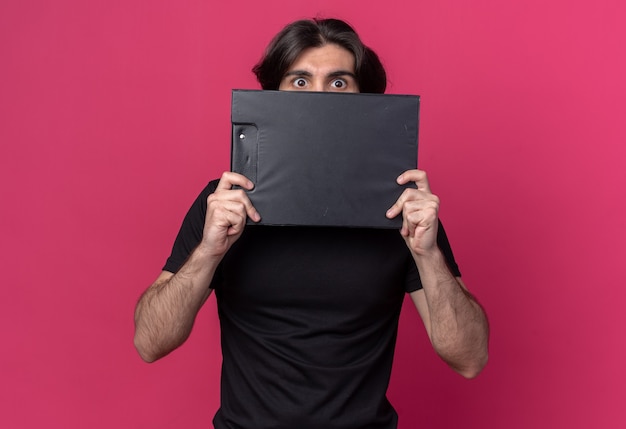 Scared young handsome guy wearing black t-shirt covered face with clipboard around face isolated on pink wall