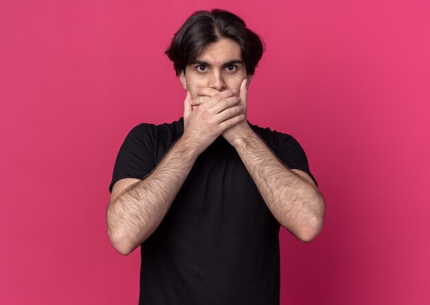 Free photo scared young handsome guy wearing black t-shirt covered mouth with hands isolated on pink wall