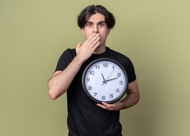 Испуганный молодой красивый парень в черной футболке держит настенные часы и прикрывает рот рукой, изолированной на оливково-зеленой стене